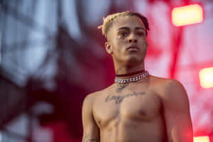 紅色舞台燈光下，金色辮子、胸前有紋身的年輕男表演者。