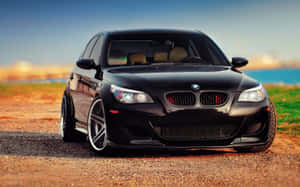 日落時分，黑色BMW M5運動轎車停泊在海岸附近的碎石路上。 — 1280x800 壁紙