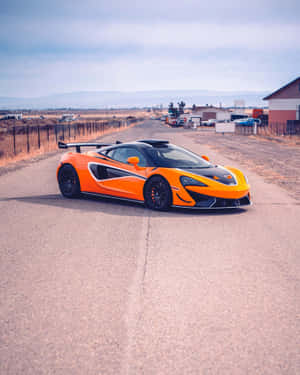橙色McLaren跑車停泊在鄉村道路上，遠處有建築物和山脈。 — Apple Watch Ultra 壁紙