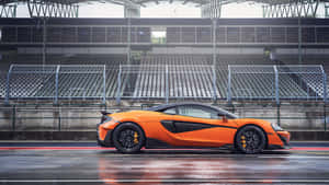 橙色McLaren跑車在潮濕的賽道上，後面是空看台。 — 5120x2880 壁紙