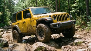 黃色Jeep Wrangler Rubicon在泥濘的森林地形上行駛，伴有岩石和飛濺的水花。