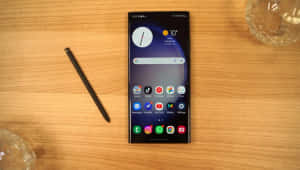 黑色智能手機和觸控筆在木桌上，旁邊有玻璃器皿 — Samsung S23 Ultra 壁紙