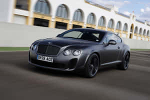 啞光灰色 Bentley Continental GT 在拱形建築附近的賽道上飛馳。 — Surface Pro 壁紙