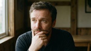 一個穿深色襯衫的鬍子男人，手托著下巴，望著窗外。