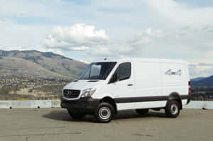 白色 Mercedes-Benz Sprinter 4x4 客貨車停泊在風景優美的觀景點，背景有丘陵和城鎮。 — Surface Laptop 壁紙