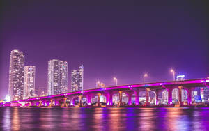 一座燈光明亮的橋樑橫跨水面，城市摩天大樓在夜晚閃爍。