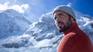 身穿紅色毛衣和灰色冷帽的男人，在日落時分站在積雪的山脈前。