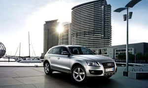 銀色 Audi Q7 SUV 停泊在海濱附近，有帆船和現代建築。 — 奧迪 GIF 壁紙