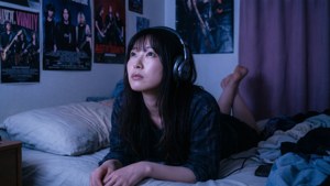 戴著耳機的年輕女子躺在昏暗房間的床上，房間有樂隊海報。
