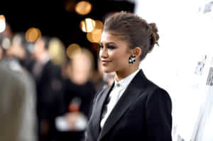 Zendaya 身穿黑色西裝在活動中微笑，身後有模糊的燈光。