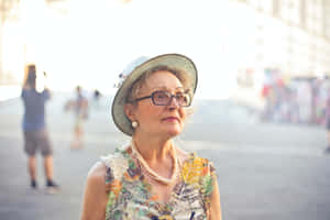 優雅的老年女士戴著眼鏡和帽子，站在陽光明媚的城市廣場上。 — 夏日風格 壁紙
