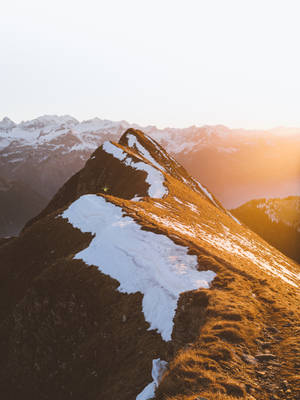 夕陽下，一位遠足者站在白雪覆蓋的山脊上，遠處有山峰。 — 旅行 壁紙