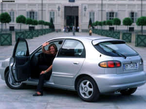 女士從宏偉建築前的銀色汽車打開車門中揮手。 — 酷車 壁紙