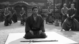 武士平靜地坐著，其他人在傳統庭院中練習劍術。 — 武士 壁紙