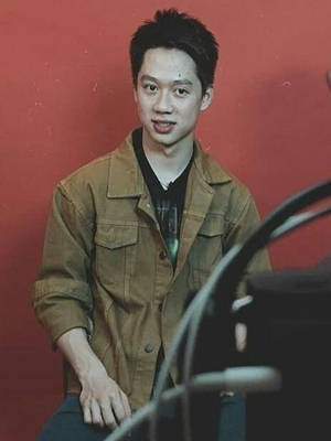 年輕男子身穿棕色外套在紅牆前微笑