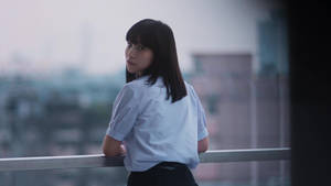 身穿藍色襯衫的年輕女子靠在陽台欄杆上眺望黃昏的城市 — Netflix GIF 壁紙