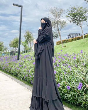 身穿黑色長袍和頭巾的女性站在公園的紫色花朵旁。 — 花系美學 壁紙