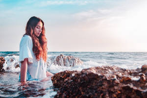 年輕女子在日落時坐在海邊岩石附近的波浪中 — 女孩 壁紙