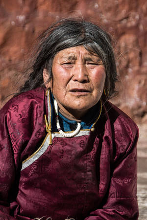 一位年長的原住民婦女，身穿傳統栗色長袍，戴著珠鍊，背景是紅色岩石。 — 中文 壁紙