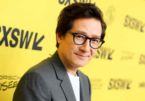 灰色西裝和眼鏡的男士在活動中對著鏡頭微笑，身後有黃色品牌牆。 — 胡桃 壁紙