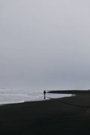 一個孤獨的人沿著黑色沙灘行走，海浪在灰色天空下翻滾。 — 海灘 壁紙