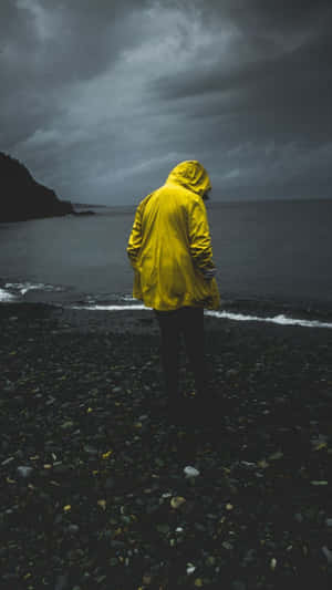 穿著黃色雨衣的人站在岩石海灘上，面對著暴風雲下的海洋 — 雨天 壁紙