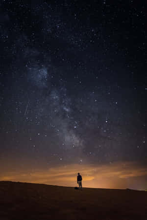 一個人在星空下的沙漠沙丘上的剪影，銀河可見。 — 暮光 壁紙