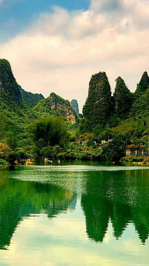 在部分多雲的天空下，平靜的湖泊中倒映著鬱鬱蔥蔥的綠色山脈。 — 向日葵田 壁紙