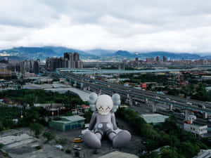 城市中大型灰色KAWS雕塑，背景有高速公路和山脈。 — 節日 GIF 壁紙