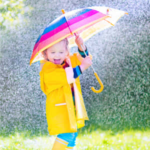 穿著黃色雨衣的孩子拿著彩虹傘在灑水器裡玩耍。 — 雨天 壁紙