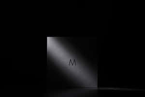 黑色正方形產品，帶有 'M' 標誌，在黑暗工作室中由聚光燈照亮。 — MacBook Pro M4 壁紙