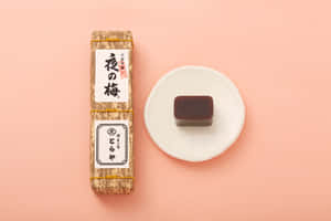 一個日本梅子醬包裝和一個深色糖果放在白色碟子上，背景是粉紅色。 — MacBook Air M3 壁紙