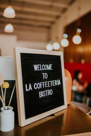 咖啡店內一個木框字母板寫著「Welcome to La Coffeeteria Bistro」，旁邊有一個白色花瓶。 — Vivo Pad 壁紙
