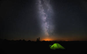 一個發光的綠色帳篷在繁星點點的夜空下，銀河橫跨山脈之上。 — 星星 壁紙