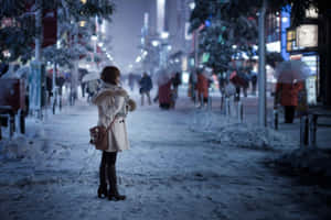 穿著白色外套的女人夜晚獨自站在積雪的城市街道上，有模糊的行人和燈光。 — 冬日美學 壁紙