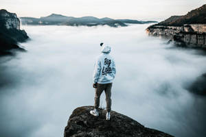 穿著白色連帽衫的人站在黃昏時分雲海之上的懸崖邊緣。 — 山脈 壁紙
