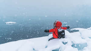 穿著紅色雪衣的小孩坐在積雪的冰上，享受著結冰湖邊飄落的雪花。 — 天使 壁紙