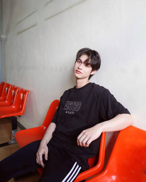 穿著黑色T恤的年輕男子坐在橙色椅子上，背景是素色牆壁 — 橙色美學 壁紙