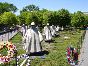 紀念公園中的灰色士兵雕像，配有色彩繽紛的花圈和綠樹，在藍天下。 — 陣亡將士紀念日 GIF 壁紙