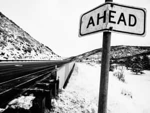 一個黑白路牌，上面寫著AHEAD，旁邊是一條積雪的高速公路，位於山區冬季景觀中。 — 黑白藝術 壁紙