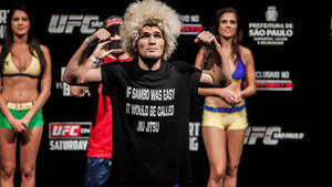 UFC拳手穿著黑色T恤在稱重時彎曲手臂，身後有比基尼模特兒。 — 動漫打鬥 壁紙