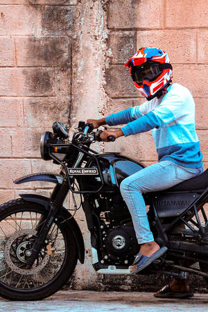 在磚牆附近，一名戴著彩色頭盔的人騎在 Royal Enfield 電單車上。 — 機車 壁紙