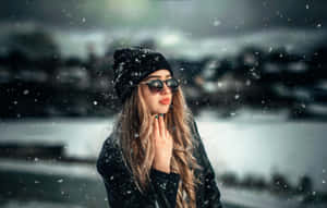 女子戴太陽眼鏡和冷帽站在水邊的雪中。 — 冬日美學 壁紙