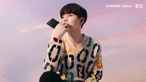 年輕男子穿著圖案毛衣，在粉彩天空下將手機貼在唇邊。 — 三星 Galaxy S20 壁紙