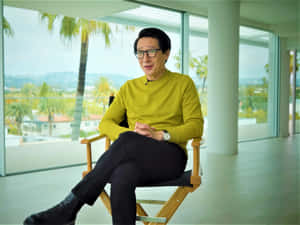 身穿黃色毛衣的男子坐在導演椅上，窗外有棕櫚樹。 — 胡桃 壁紙