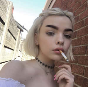 金髮女性穿著露肩上衣，靠著磚牆抽煙。