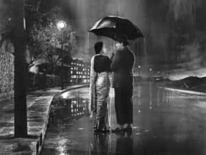 情侶在雨夜的街道上共享雨傘，伴隨著閃爍的路燈。 — 懷舊美學 壁紙
