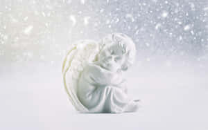 在冬日場景中，一座白色天使雕像在飄落的雪花下蜷縮入睡。 — 2880x1800 壁紙