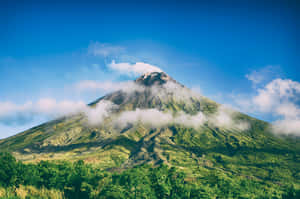 在明亮的藍天下，一座鬱鬱蔥蔥的綠色火山，雲霧繚繞其峰頂。 — MacBook Pro 16 壁紙