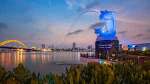 藍色龍雕像在暮色中向河流噴水，城市燈光閃爍。 — 地龍 壁紙